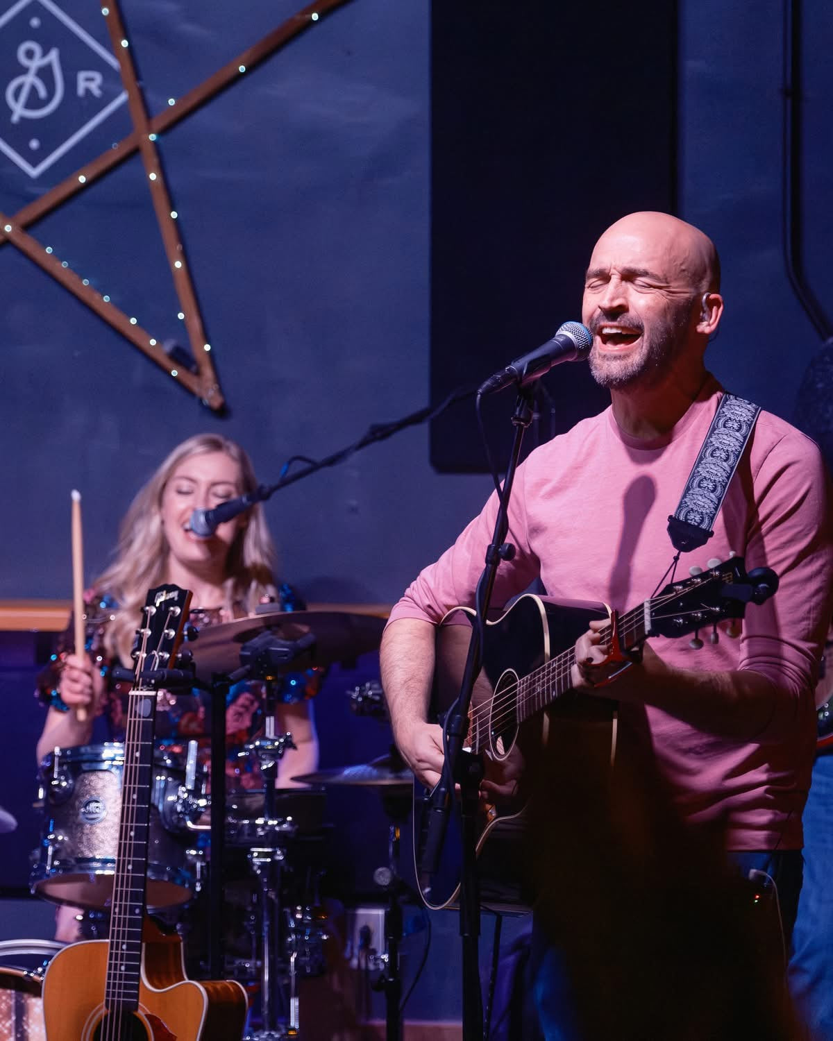 Man playing guitar and singing into a microphone on stage with a woman playing drums in the background.