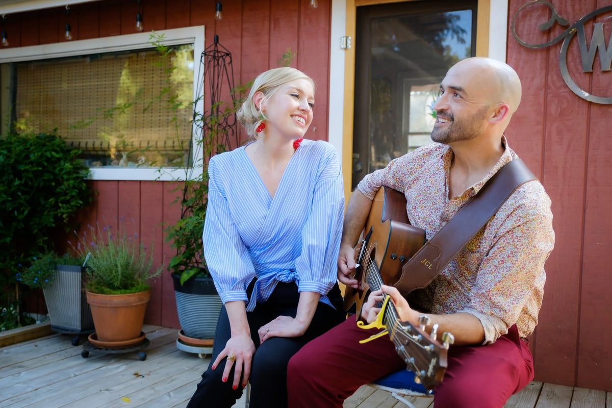 Man playing guitar and woman sitting next to him in front of a red building.